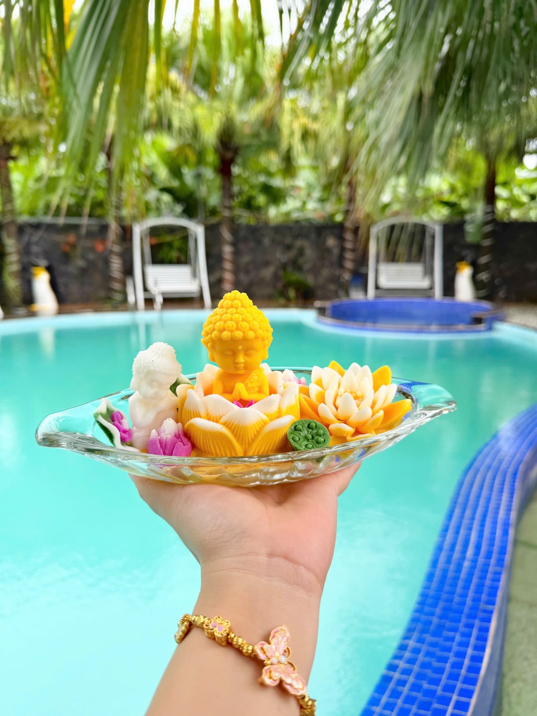 Bàn tay nâng đĩa thuỷ tinh có tượng Phật sáp vàng ngồi trên đài sen, quanh là sen vàng và gương sen