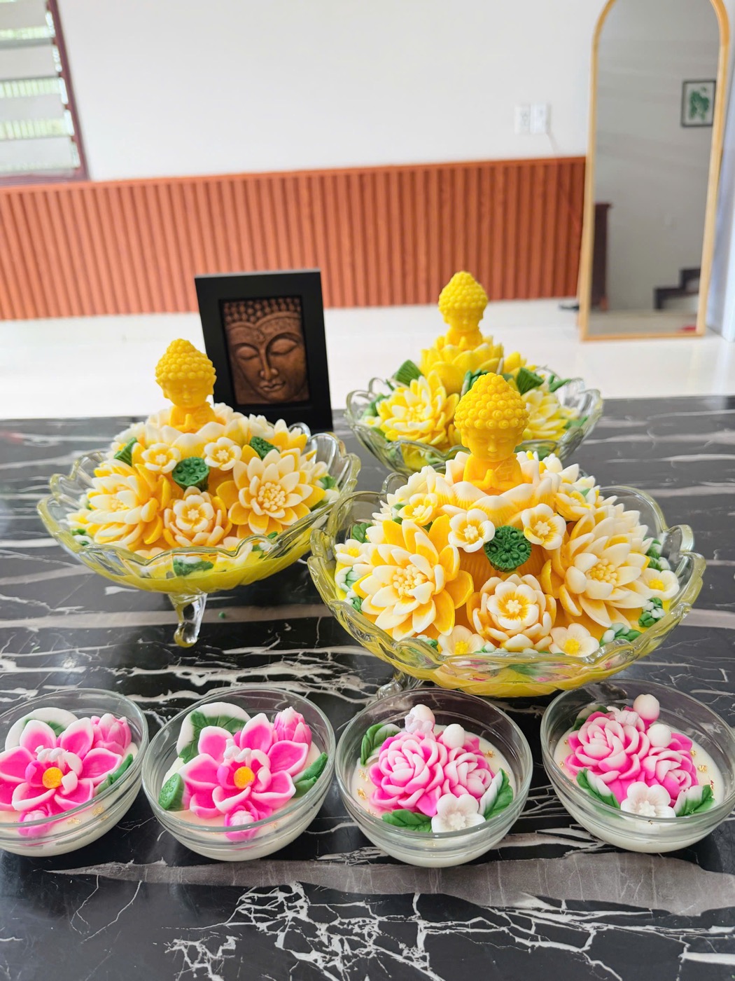 Ba tô Phật hoa sen sáp vàng đặt cạnh bốn bát nến hoa hồng trên mặt bàn đá — hàng dâng lễ của Nến Hoa Khuê Các